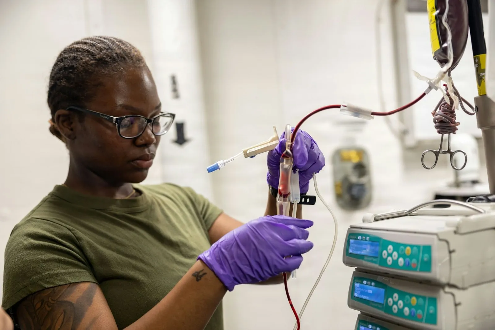 A clinician in gloves checking a line beside a stack of infusion pumps